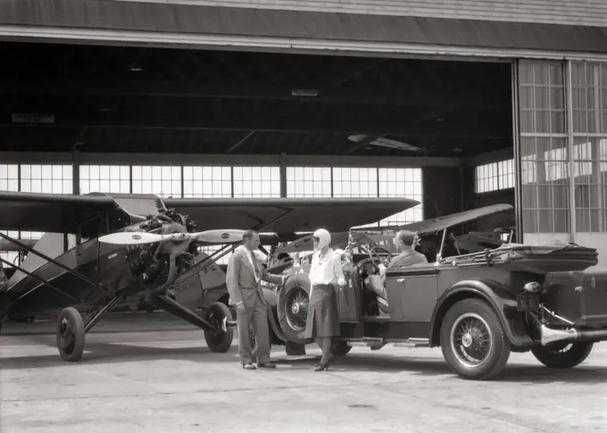 1920s 1930s COUPLE WITH CONVERTIBLE PACKARD CAR TALKING TO AIRPLANE SALESMAN AT HANGAR 