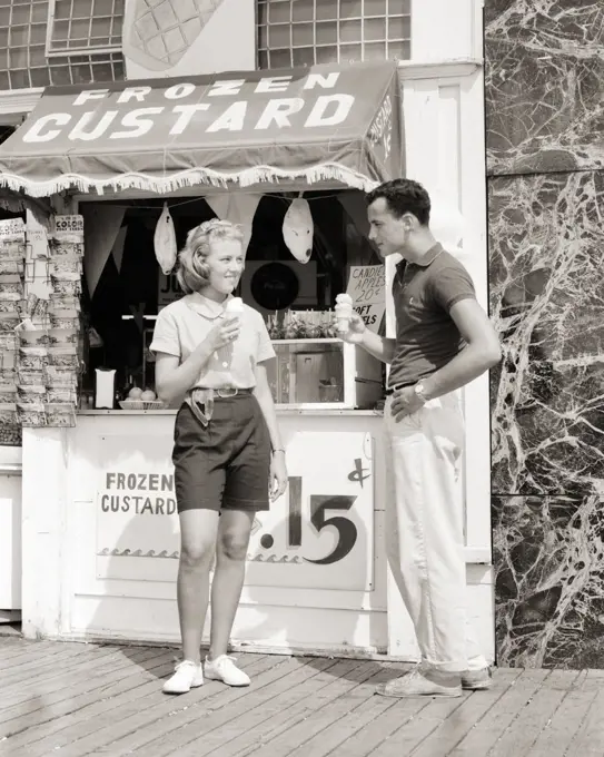 1950s 1960s DATING TEENAGE COUPLE EATING FROZEN CUSTARD CONES STANDING IN FRONT OF SEASHORE BOARDWALK STAND NEW JERSEY USA