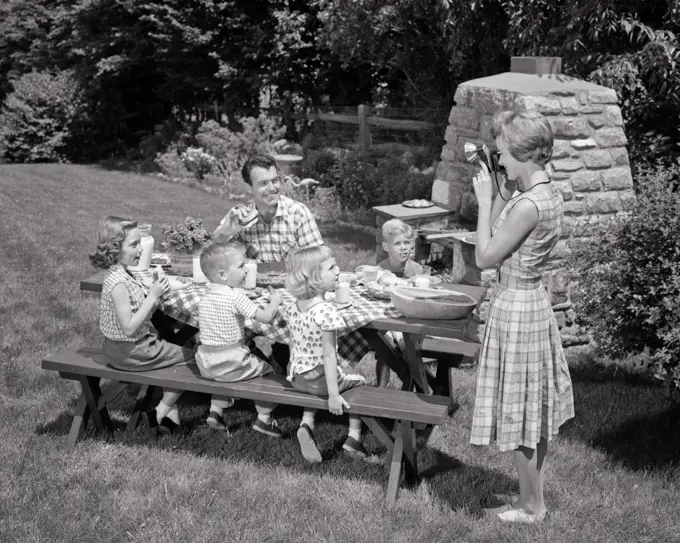 1960s MOTHER TAKING SNAPSHOT PHOTO OF FAMILY PICNIC BACKYARD BBQ KIDS AND DAD AT TABLE 