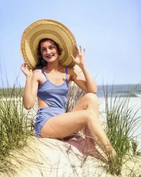 1920s 1930 SMILING BATHING BEAUTY WEARING STRAW HAT SITTING ON BEACH SAND DUNE LOOKING AT CAMERA