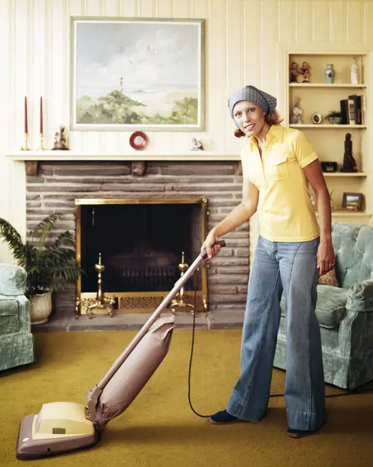 1970s WOMAN HOUSEWIFE WEARING BELL-BOTTOM JEANS YELLOW T-SHIRT HEADSCARF VACUUMING LIVING ROOM USING UPRIGHT VACUUM