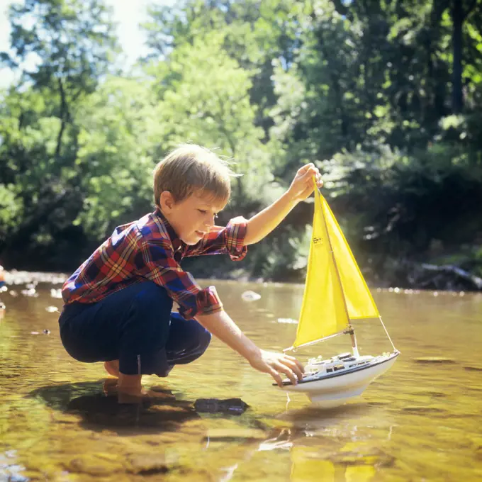 1970s BOY ABOUT TO LAUNCH A TOY SAIL BOAT INTO A STREAM