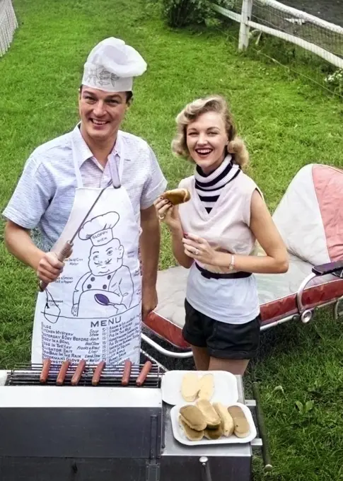 1950s 1960s COUPLE BACKYARD GRILLING LOOKING AT CAMERA COOKING HOT DOGS MAN WEARING APRON TOQUE & SKEWERED HOT DOG