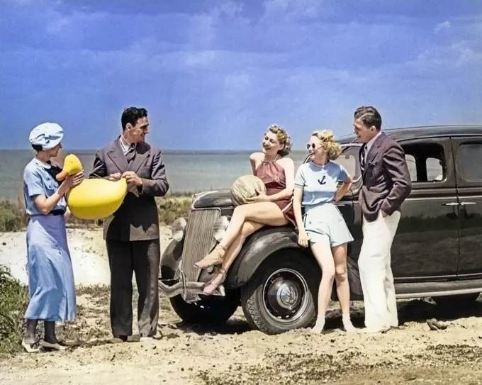 1930s FIVE MEN & WOMEN IN CASUAL SUMMER CLOTHES PLAYING WITH INFLATABLE BEACH TOYS BY 1936 FORD V-8 SEDAN PARKED ALONG SEA SHORE