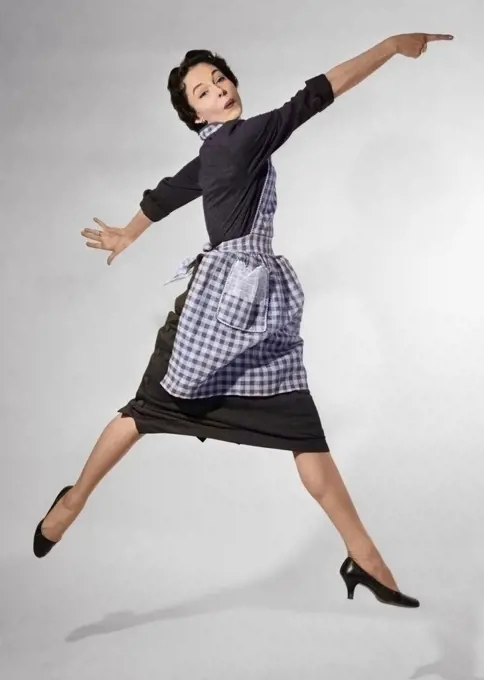 1950s WOMAN HOUSEWIFE IN GINGHAM APRON RUNNING AND JUMPING INTO AIR SMILING AND POINTING INDEX FINGER LOOKING AT CAMERA