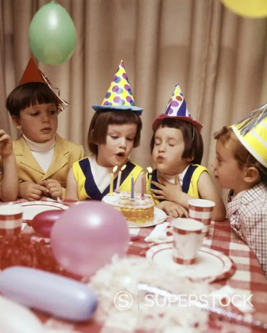 1970s TWIN GIRLS AND OTHER CHILDREN AT BIRTHDAY PARTY BLOWING OUT CANDLES ON CAKE