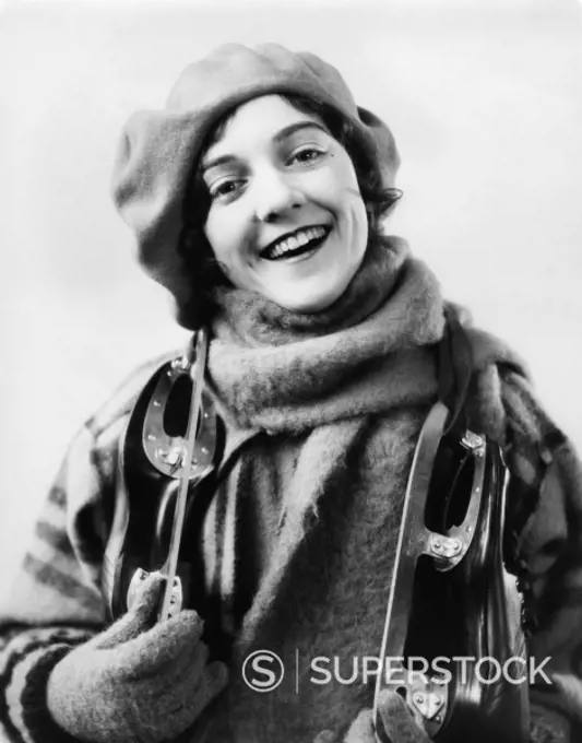 1920s 1930s SMILING WOMAN DRESSED FOR WINTER LOOKING AT CAMERA PAIR OF ICE SKATES OVER HER SHOULDERS