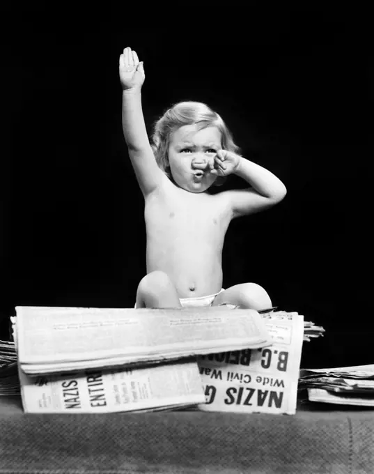 1940s LITTLE BLOND TODDLER GIRL IMITATING HITLER GIVING NAZI SALUTE SITTING ON PILE OF NEWSPAPERS