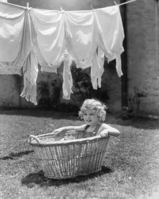 1930s 1940s GIRL OUTDOORS SITTING IN LAUNDRY BASKET UNDER CLOTHESLINE FULL OF SHIRTS