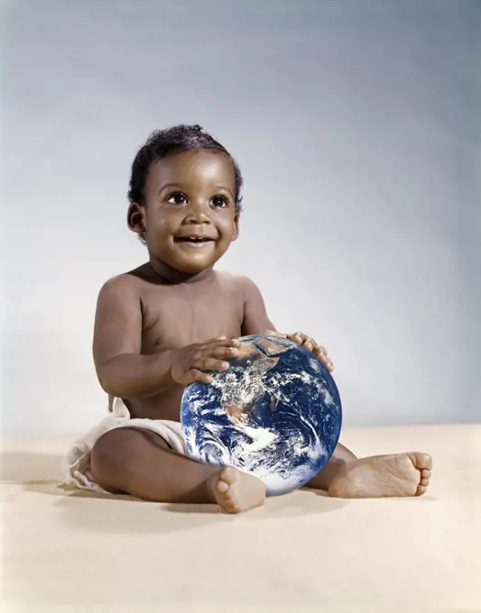 1960s SYMBOLIC ECOLOGY SMILING AFRICAN AMERICAN BABY WEARING CLOTH DIAPERS SITTING HOLDING THE EARTH LOOKING TO THE FUTURE