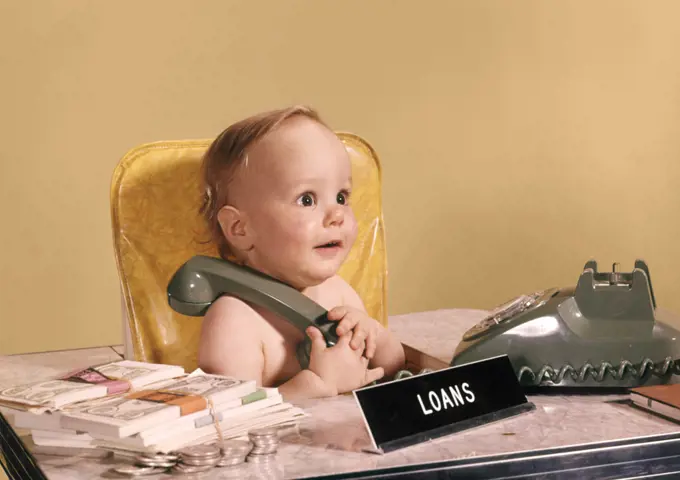 1960s WIDE EYED BABY SEATED AT LOAN OFFICER DESK HOLDING TELEPHONE
