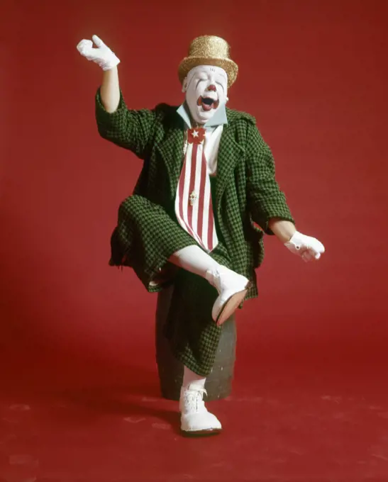 1970s CLOWN GREEN CHECKED BAGGY SUIT SILLY EXPRESSION SITTING ON BARREL LIFTING LEG ARMS IN AIR YAWNING