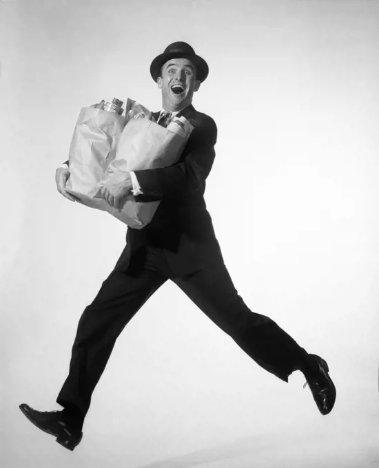 1950s 1960s HAPPY EXCITED MAN WEARING BUSINESS SUIT HAT CARRYING GROCERY SHOPPING BAGS RUNNING JUMPING IN AIR LOOKING AT CAMERA