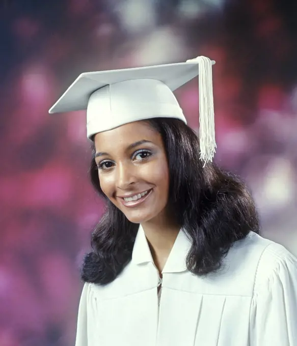 1970s YOUNG AFRICAN AMERICAN WOMAN GRADUATE IN WHITE CAP AND GOWN LOOKING AT CAMERA