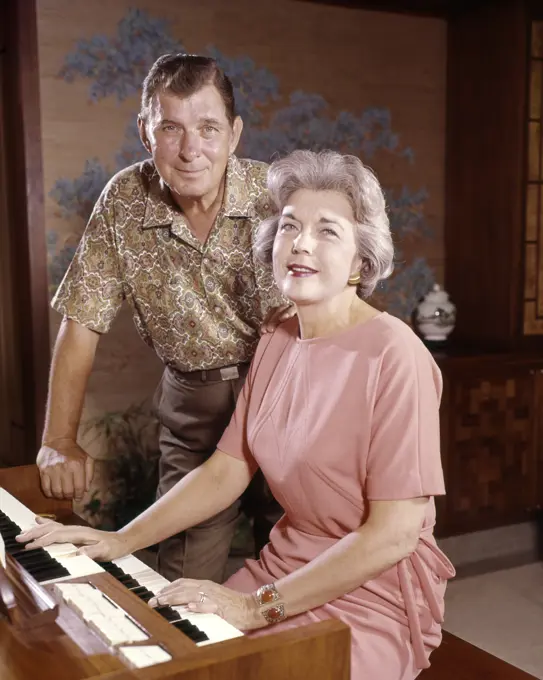 1960S SMILING SENIOR COUPLE TOGETHER AT ELECTRIC HOME ORGAN HUSBAND LOOKING AT CAMERA STANDING BEHIND WIFE WHO IS PLAYING