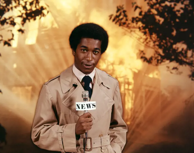 1970s AFRICAN AMERICAN TV NEWSMAN WEARING TRENCH COAT LOOKING AT CAMERA HOLD MICROPHONE REPORTING AT SCENE OF RAGING HOUSE FIRE