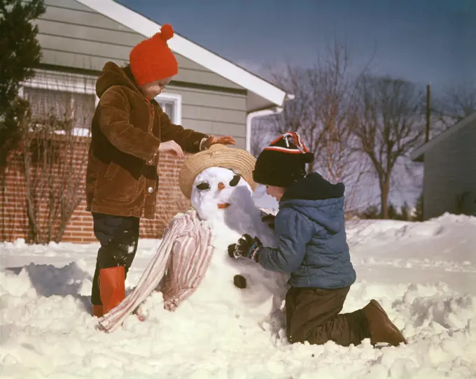 1960s 1970s BOY AND GIRL BROTHER AND SISTER MAKING SNOW MAN IN SUBURBAN HOUSE FRONT YARD