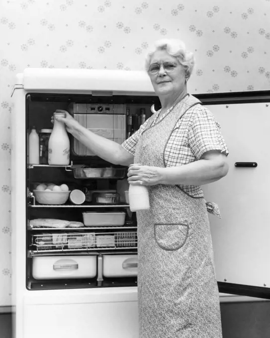 1930s 1940s SENIOR WOMAN HOUSEWIFE GRANDMOTHER LOOKING AT CAMERA TAKING TWO GLASS BOTTLES OF MILK OUT OF KITCHEN REFRIGERATOR