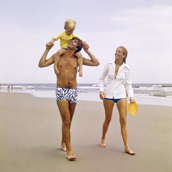 1970s YOUNG FAMILY WALKING DOWN BEACH TODDLER RIDING ON FATHERS SHOULDERS
