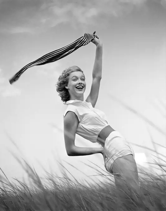 1950S 1960S Smiling Young Woman In White Shorts And Blouse Bending Back Waving Scarf In The Wind Standing On Grassy Beach Dune