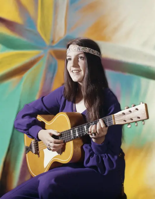 1970S Young Woman Wearing Purple Dress And Beaded Headband Playing Acoustic Guitar Patterned Background Folk Song