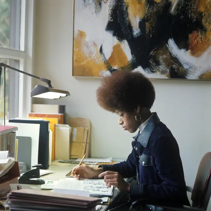 1960S 1970S African American Woman Student Sitting At Desk Writing Papers By Window Light Afro Hair Style Denim Jacket
