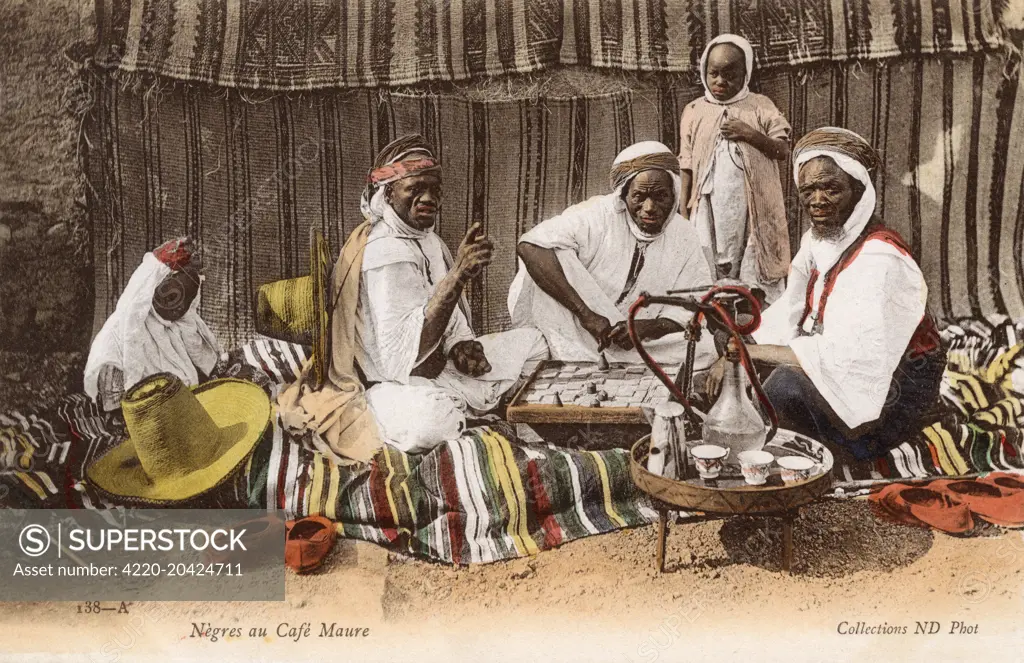 Black African men taking tea and a smoke of a hookah pipe at a drink at a Moorish Cafe - Algeria. Two of the men are playing a board game.     Date: circa 1910s