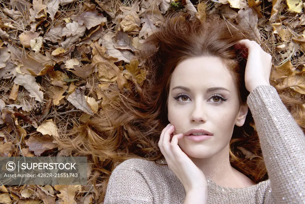 An attractive young woman laying on the forest floor surrounded by leaves.