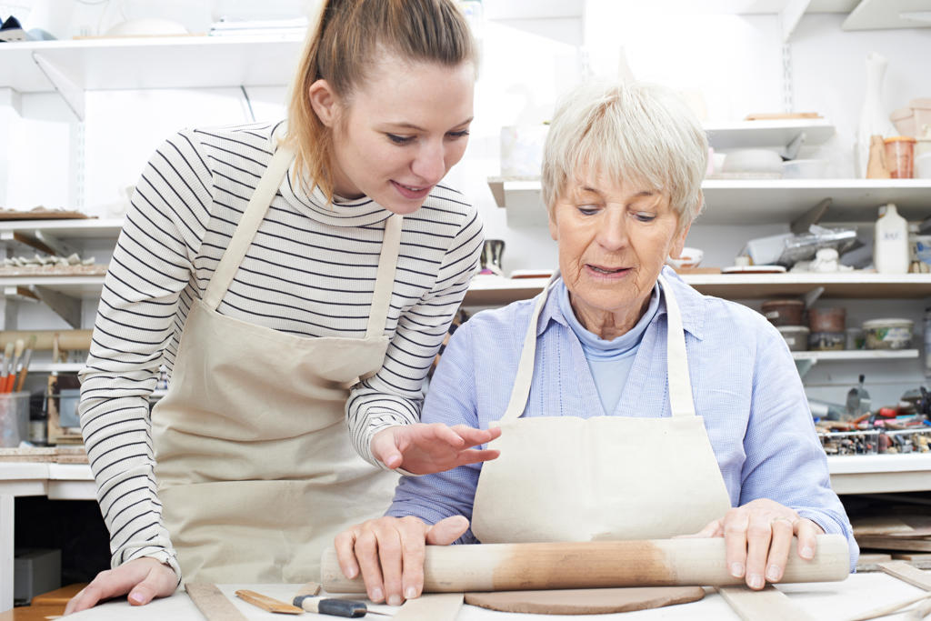 Senior Woman Rolling Out Clay With Teacher In Pottery Class