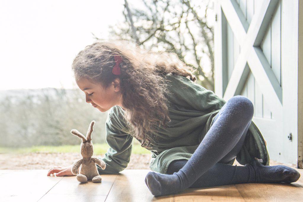 A six year old girl sitting at a back door playing with a toy rabbit.