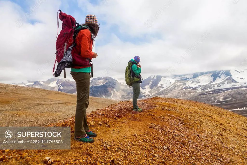 Two women backpacking and taking in the view of Mount Katmai from the rainbow-coloured pumice, rock and clay slopes of Broken Mountain, Valley of Ten ...