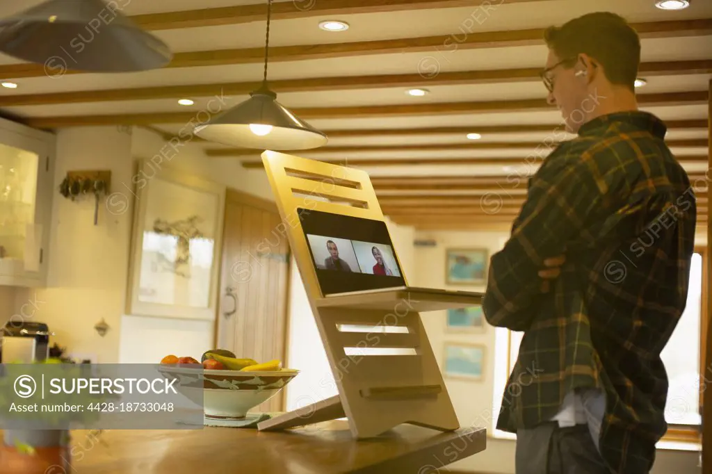 Man video conferencing with colleagues at laptop stand desk in kitchen