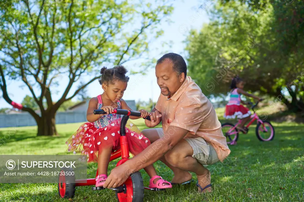 Grandfather helping granddaughter ride tricycle in summer park