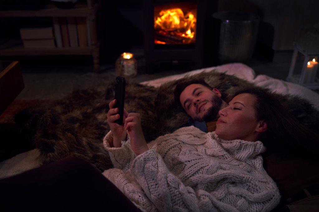Affectionate couple using smart phone on blankets at cozy fireside