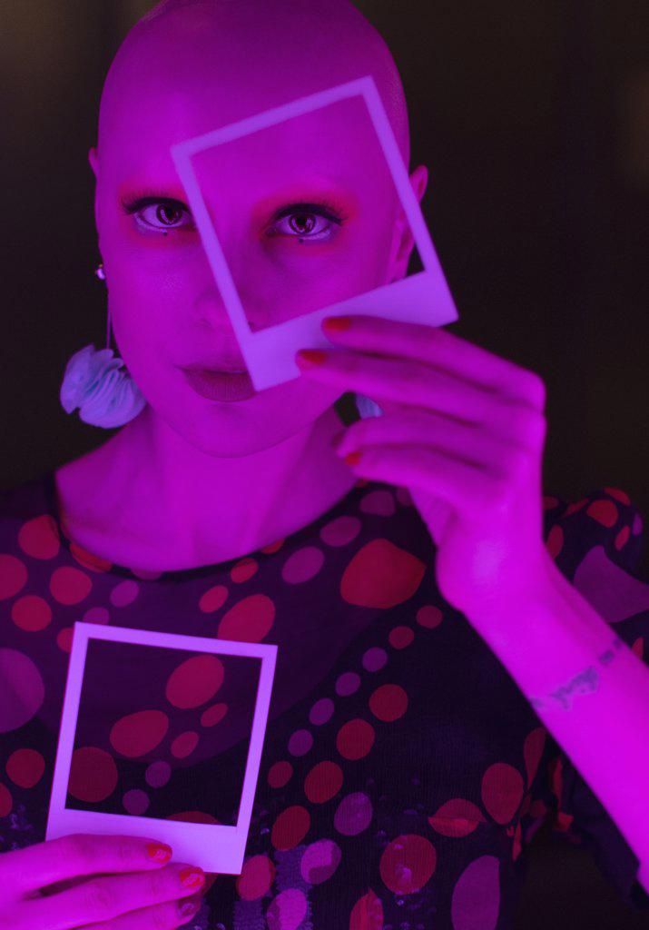 Portrait stylish woman with shaved head holding polaroid over eye