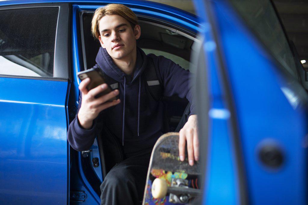 Teenage boy with skateboard using smart phone in car doorway