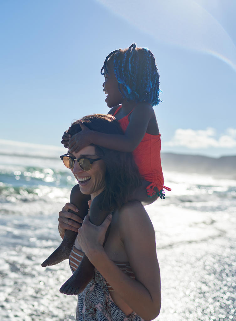 Happy mother carrying cute daughter on shoulders on sunny ocean beach