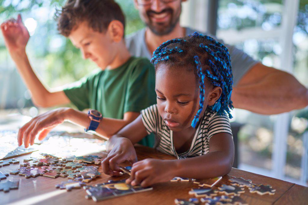 Cute toddler girl assembling jigsaw puzzle with family at table