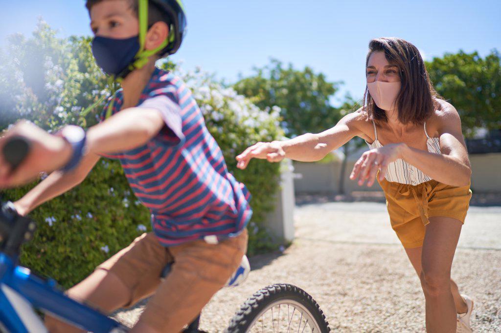 Mother in face mask pushing son on bicycle in sunny driveway