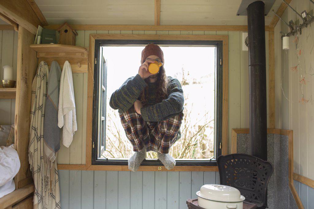 Young man in pajamas drinking coffee in tiny cabin window