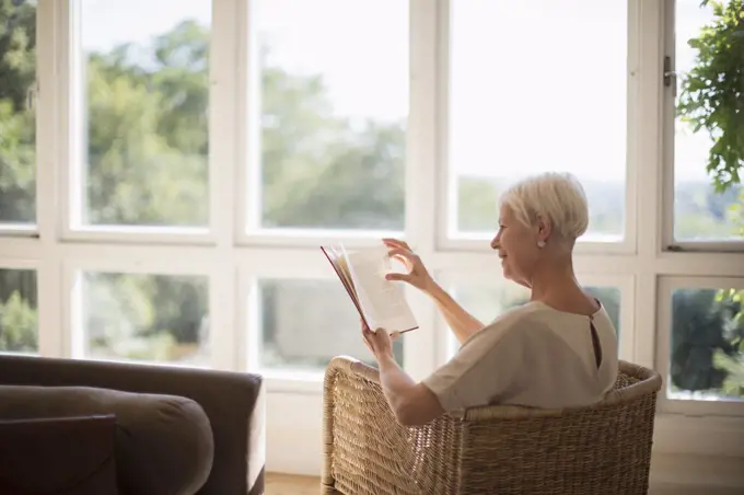 Senior woman relaxing and reading book in sunny living room
