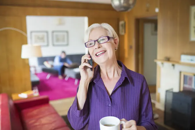 Happy senior woman with coffee talking on smart phone in living room