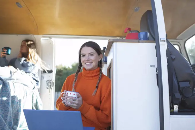 Portrait happy young woman with coffee using laptop in camper van