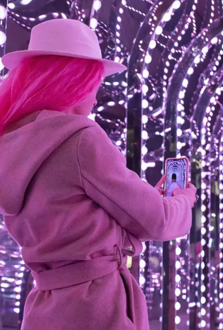 Fashionable woman in pink coat using camera phone under lights