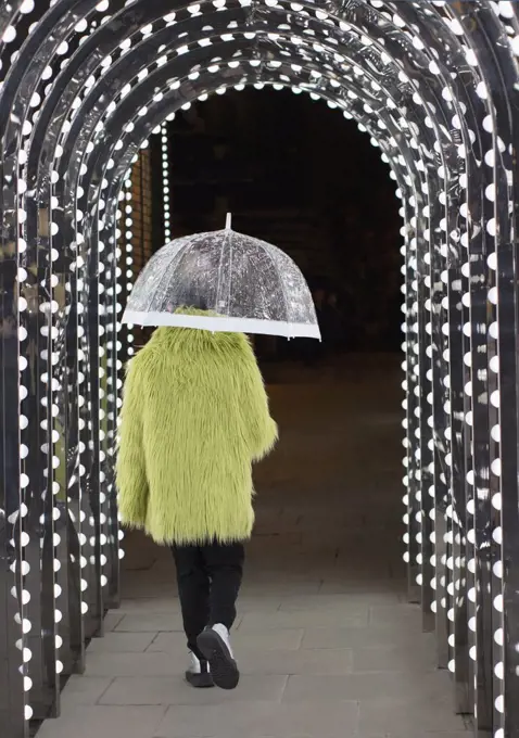 Eccentric young man in feather coat with umbrella under arch lights