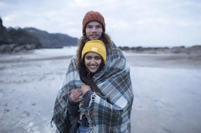 Portrait happy young couple wrapped in blanket on cold beach