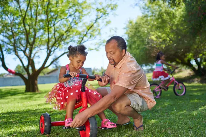 Grandfather helping granddaughter ride tricycle in summer park