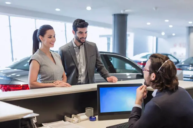 Couple customers talking to male receptionist at desk in car dealership showroom