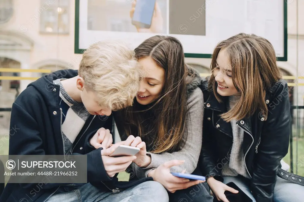 Smiling friends using mobile phone while sitting at bus stop