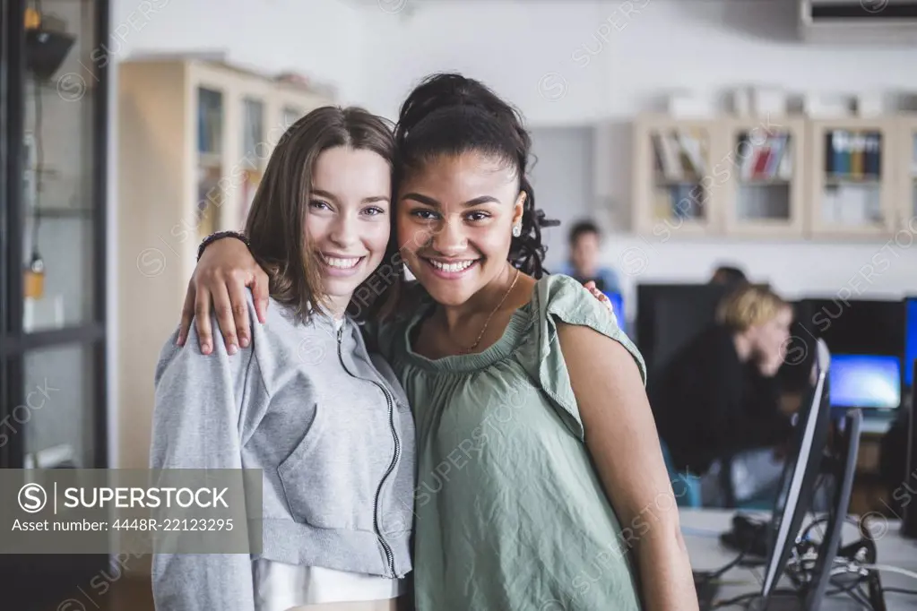 Portrait of smiling female teenage friends standing with arm around in computer lab at high school
